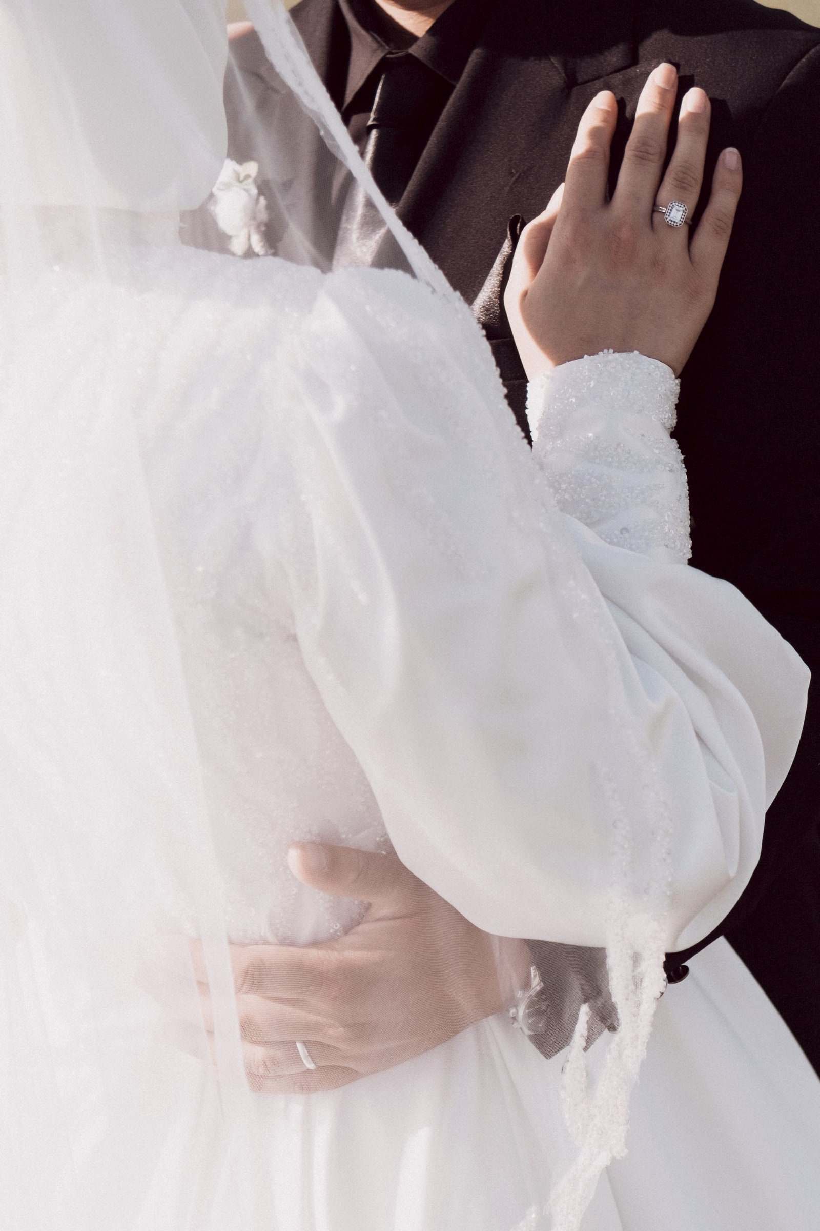 A groom's hand with a wedding ring resting on the bride's back, her veil over her shoulder