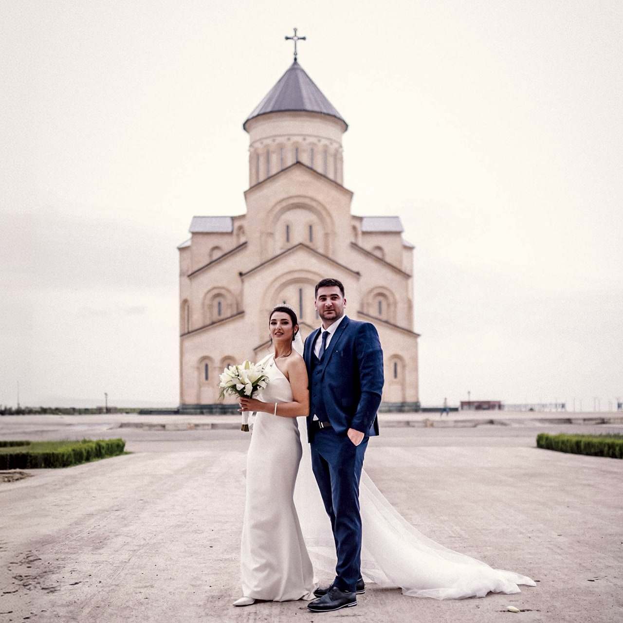 A bride with a bouquet and a groom in a blue suit in front of the Holy Trinity Cathedral in Tbilisi