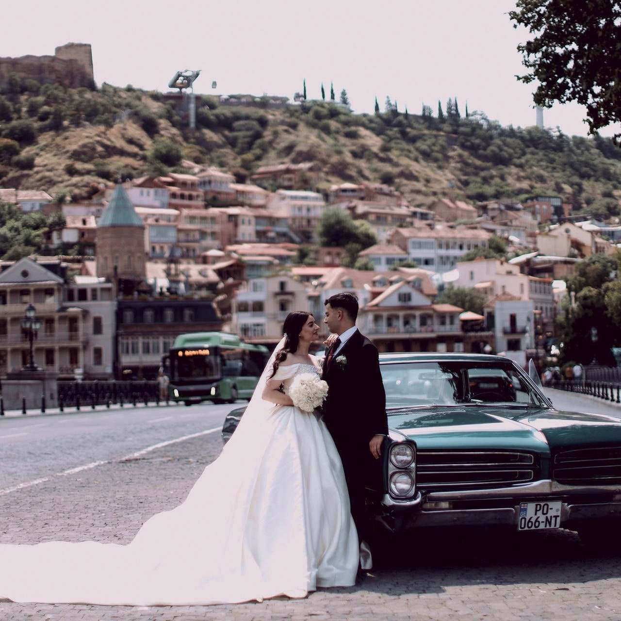 A bride and groom beside a vintage green car in the old town of Tbilisi, the fortress on the hill behind