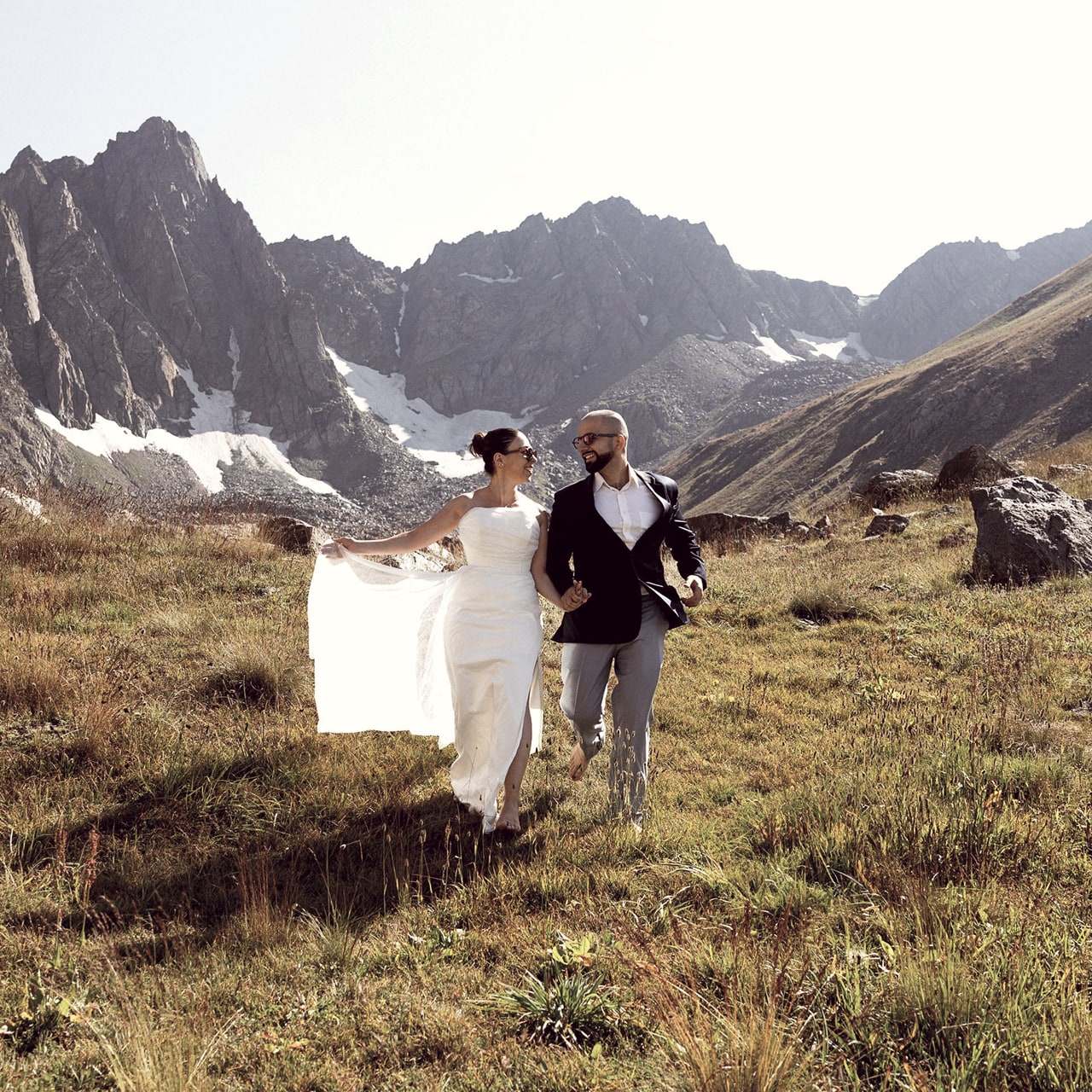 A bride and groom walking hand in hand through an alpine meadow beneath snow-capped peaks in the Caucasus